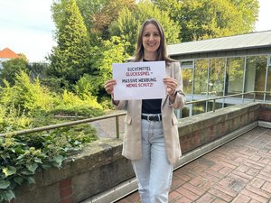 Polina Lehmann, Fachkraft für Glücksspielprävention und -beratung bei der Diakonie, hält ein Schild mit der Aufschrift ‚Suchtmittel Glücksspiel – Massive Werbung. Wenig Schutz!‘ in die Kamera.